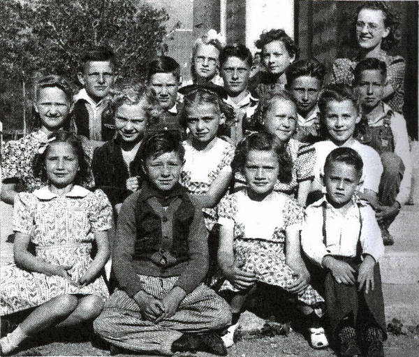 A group of seventeen children, both boys and girls, are seated and standing in rows outside, likely in front of a school building. Most of the children are wearing dresses or shirts with collars, and some have bows in their hair. An adult stands behind the group, smiling. Trees and part of a building are visible in the background.