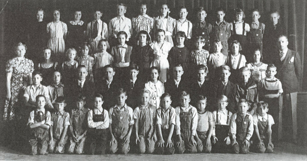 A large group of children, mostly boys, is arranged in multiple rows, with some adults standing on either side. They are wearing a variety of clothing, including overalls and dresses. The setting appears to be indoors with a curtain in the background, suggesting a formal group gathering such as a school class photo.