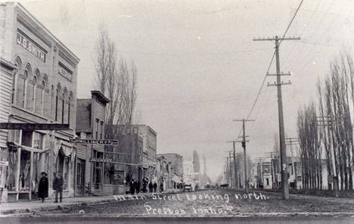 A street scene with buildings lining both sides, featuring businesses such as J.D. Smith and others with awnings. Several people are walking along the sidewalk. There are tall trees and utility poles on the right side of the street. Text at the bottom reads: "Main Street Looking North, Prosser, 1910s."