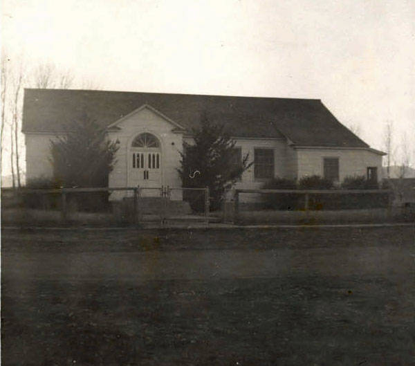 A single-story building with a gabled roof and an arched window above the entrance. The entrance is flanked by shrubs and is set back from a fenced area, fronted by a dirt road.