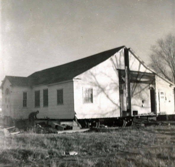 A building on a flat surface, possibly being relocated. It's a small structure with a gabled roof and several windows. There is open space around the building with grassy ground and scattered debris. The shadow of a tree is visible on the side of the building.