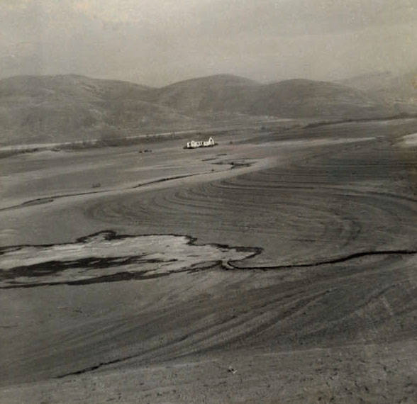 A solitary building is situated in the middle of a wide, flat expanse with swirling patterns on the ground. The landscape is surrounded by distant, rolling hills.