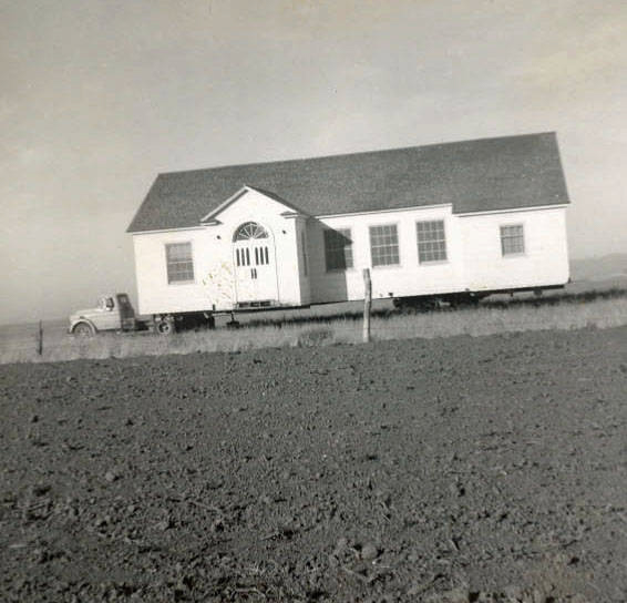 A large house is being transported on a flatbed truck across a plowed field. The house has a prominent front door with an arched window above it and several windows along the side. The surrounding area is open land.