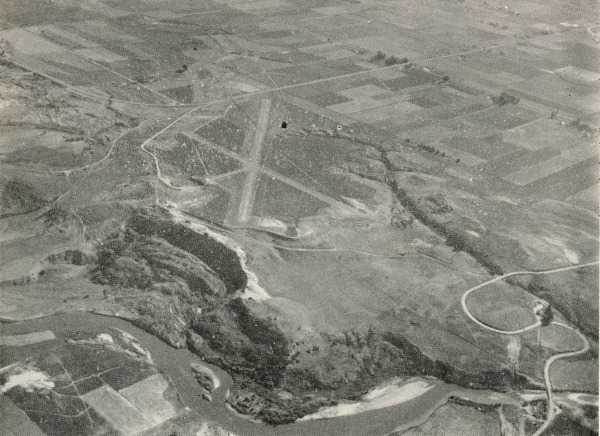 Aerial view of a landscape featuring a runway, several winding roads, and a river. The surrounding area includes a patchwork of fields and open land. The scene is expansive, with no text visible.