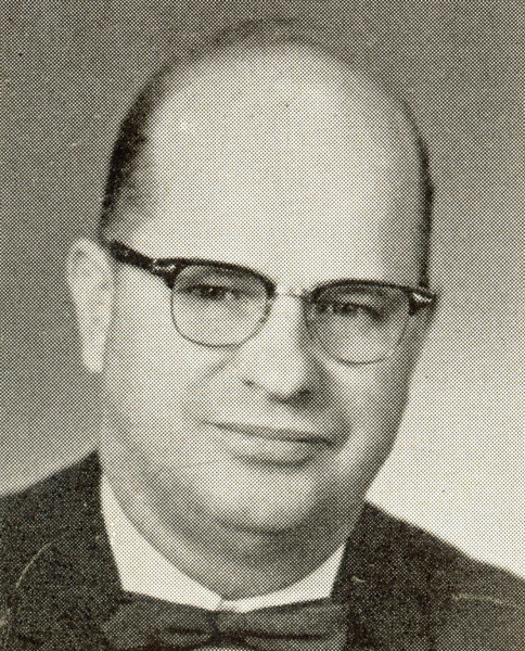 A man with glasses and a bow tie is looking at the camera. He has a bald head and is wearing a collared shirt.
