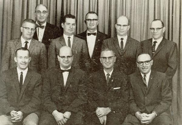 A group of ten men in suits pose together in front of a curtain. Some are wearing glasses and most have ties. The men are arranged in three rows, with four standing in the back, three in the middle, and three seated in the front.