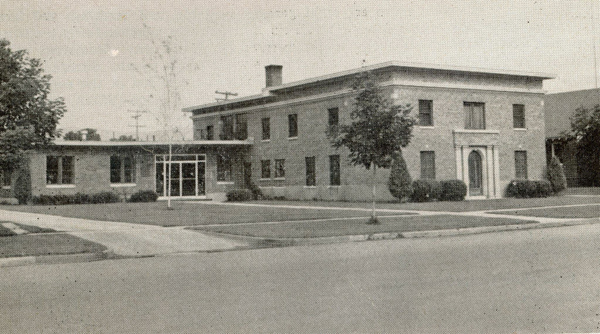 A two-story brick building with a rectangular structure, surrounded by neatly trimmed shrubs and small trees. The entrance features a decorative arch above the doorway. The building is set back from the street with a lawn and sidewalks leading up to it. There are power lines visible in the background.