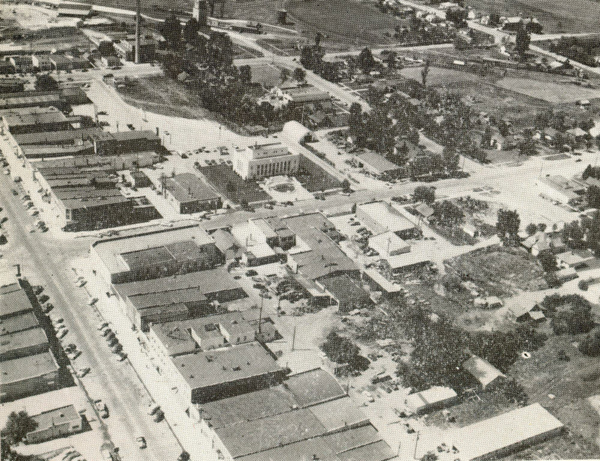 An aerial view of a small town with a grid layout of streets and buildings. There are several one-story structures, likely shops or houses, and a few parked cars along the streets. Trees and small patches of greenery are scattered between the buildings. There is a larger, more prominent building near the center, possibly a public or municipal structure. In the background, expansive open land is visible.