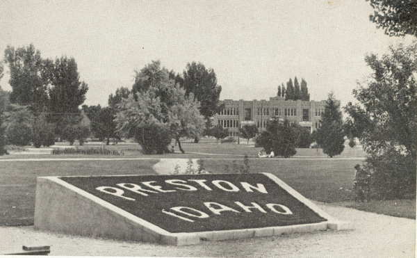A landscaped area with a prominent sign displaying the words "PRESTON IDAHO." In the background, there is a large building with numerous windows, surrounded by trees and pathways.