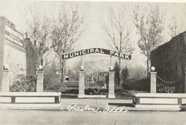 A decorative iron gate with stone pillars stands in front of a park. A sign above the entrance reads "Municipal Park." There are trees in the background. The word "Cedar City" appears at the bottom.