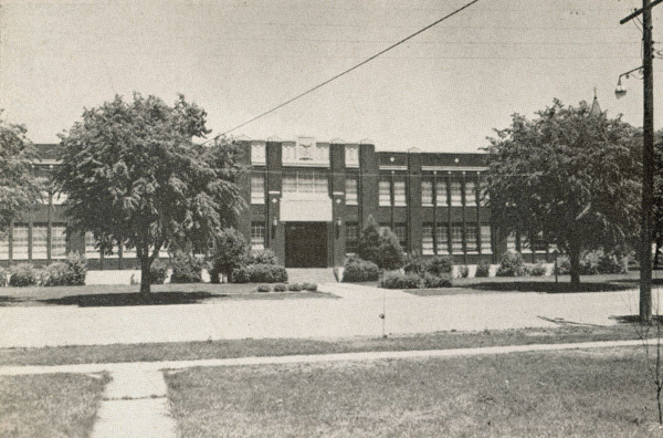 A large brick building with two rows of windows and a central entrance. Trees and shrubs are in front of the building. A sidewalk leads to the entrance, and there is a telephone pole and power line in the foreground.