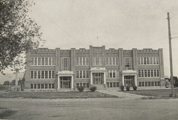 A large three-story brick building with a symmetrical facade and numerous windows. The entrance is centered with steps leading up to it, flanked by columns. The building is set back from the street by a lawn, with a tree on the left side and a utility pole on the right. There are two pathways leading from the street to the entrance.