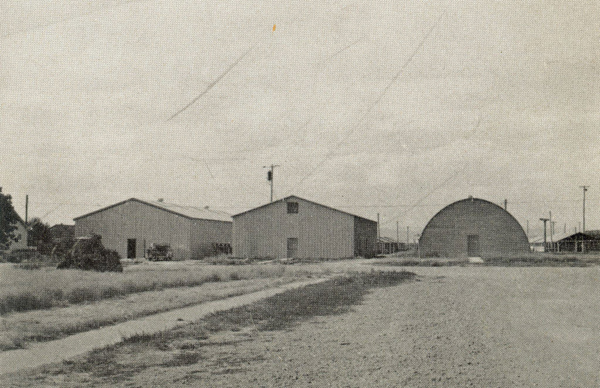 A landscape with three large buildings. The first two are rectangular with pitched roofs, and the third has a rounded, Quonset hut shape. The buildings are surrounded by open, grassy fields with a dirt path in the foreground. Power poles and a few scattered objects are visible around the buildings.