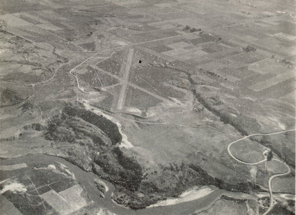 Aerial view of a landscape showing a large expanse of farmland divided into rectangular plots. A river winds through the lower part of the scene. There are visible roads and a runway crossing the fields. Sparse vegetation and gentle hills are scattered throughout the area.