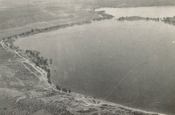 An aerial view of a large body of water with a narrow road running parallel to the shoreline. The landscape is dotted with trees and the water extends to the horizon, surrounded by open land.