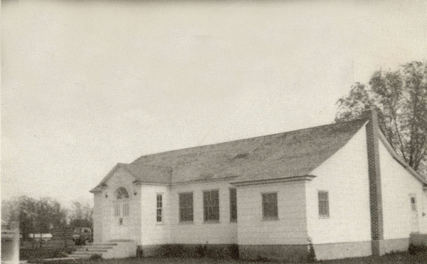 A small, single-story building with a sloped roof and arched windows. It has a chimney on one side. There are trees and a vehicle in the background, and a staircase leads to the entrance on the left side.