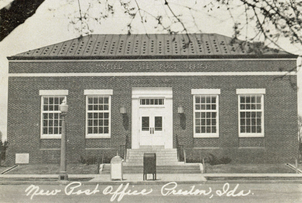A brick building with a flat roof and a symmetrical facade featuring a central entrance flanked by large windows. The words "UNITED STATES POST OFFICE" are written above the entrance. A lamppost and a mailbox are situated in front of the building. The text "New Post Office Preston, Ida." is written across the bottom. Tree branches are visible in the foreground.