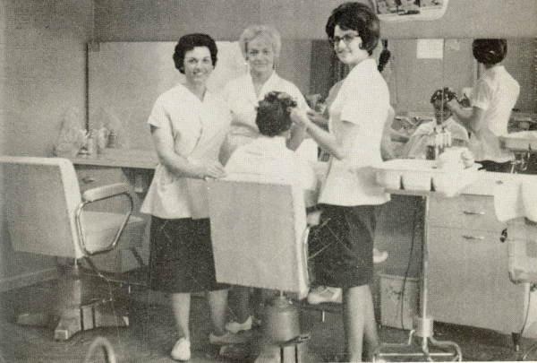 Three women in uniform are standing around a seated person in a beauty salon. One is styling the seated person's hair, while the others are observing. The salon has styling chairs, mirrors, and various hair products and tools on the counter. Another stylist is visible in the background working on a client.