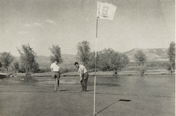 Two men are golfing on a course with a flag labeled "19" in the foreground. One man is putting while the other watches. Trees and mountains are visible in the background.