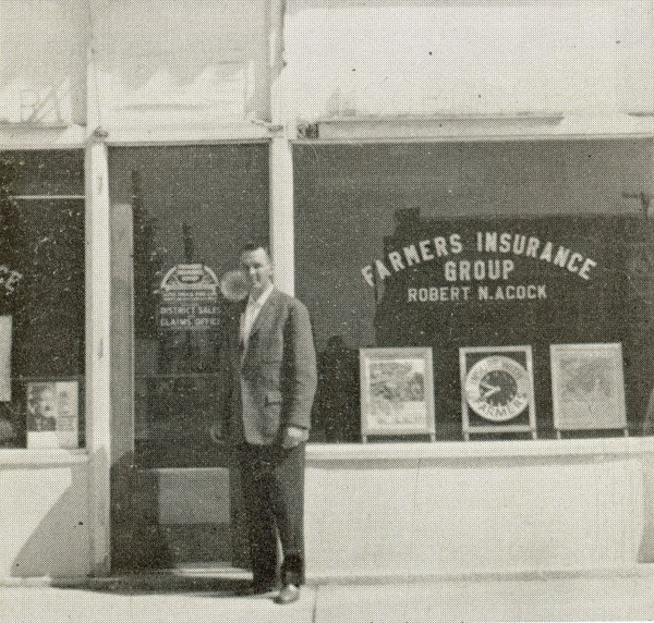 A man in a suit stands in front of a storefront. The window displays the text "Farmers Insurance Group Robert W. Acock." Signs and posters are visible inside the window.