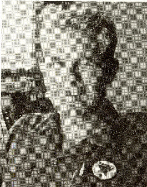 A man with short, light hair is smiling. He is wearing a buttoned shirt with a round pin featuring a design on the left side. There are shelves with books behind him.