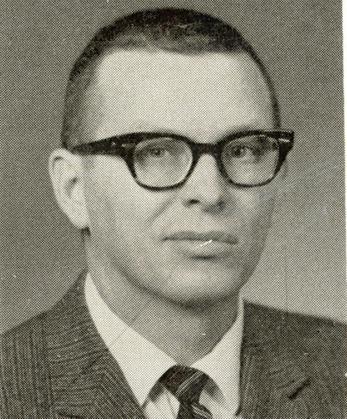 A man with short hair and glasses, wearing a suit and tie, is facing forward with a neutral expression.