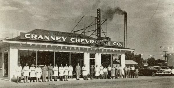 A group of people stands in front of a building with large windows. The sign on the building reads "CRANNEY CHEVROLET CO." An additional sign above reads "CHEVROLET." Two cars are parked to the right of the group. Behind the building, there is a tall chimney emitting smoke.