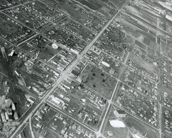 An aerial view of a city layout showing a grid of streets with various small buildings and houses. There are patches of open land and fields. The intersections are clearly visible, and there are some larger buildings interspersed among smaller structures.
