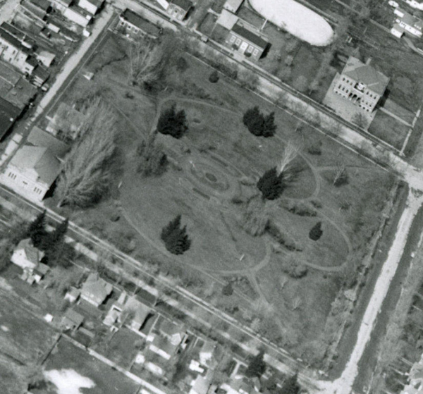 Aerial view of a rectangular park with curved pathways and several trees spread throughout. The park is surrounded by roads and residential buildings. Some larger structures are visible on the perimeter, including a distinctive rectangular building and an oval-shaped area.