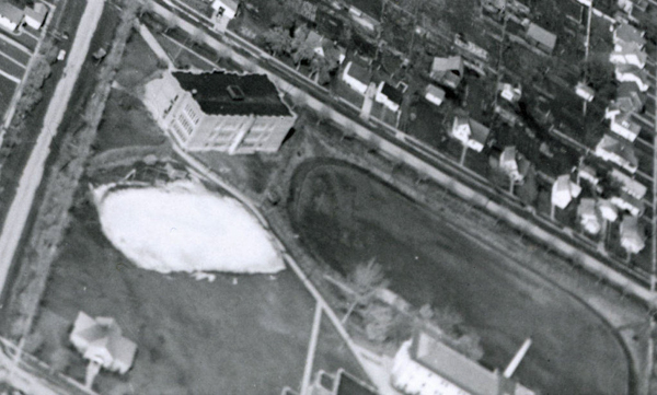 An aerial view of a landscape featuring a large rectangular building and an adjacent oval-shaped area. To the right, a series of small residential houses are lined along a street. There are various roads and patches of grassy areas surrounding the structures.