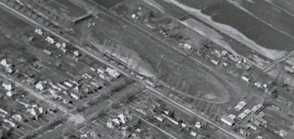 An aerial view of a neighborhood with rows of small houses lined along streets. There is an oval-shaped area that appears to be a track or park surrounded by more houses. Fields can be seen in the background.