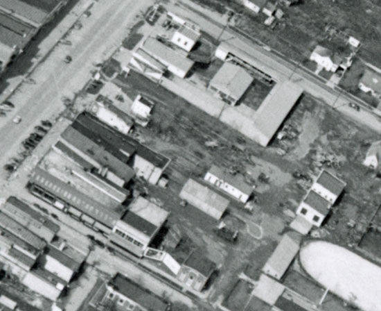 An aerial view shows a city block with multiple buildings of varying sizes. There is a main road visible on the left side with several parked cars along it. The buildings are arranged in a grid-like pattern with open spaces in between. A large circular shape is visible in the lower right corner, possibly a tank or pool. The area includes a mix of commercial and residential structures, with some open lots and pathways visible.