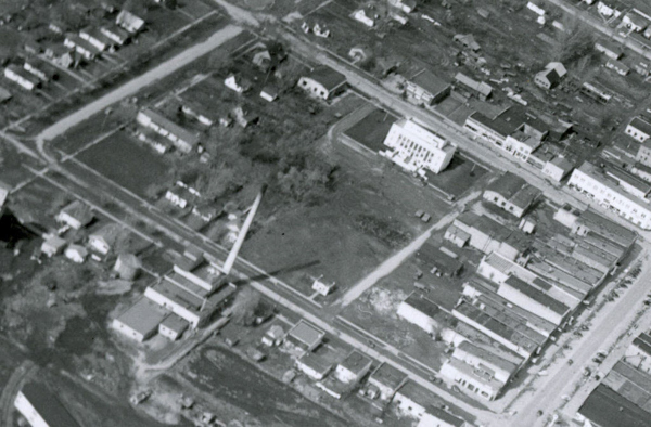 An aerial view of a town featuring grid-like streets lined with small buildings. A larger, prominent building with a front staircase is situated within a spacious yard area. Roads and intersections divide residential areas, with scattered houses and open plots visible. There are several small structures and trees dispersed throughout the scene.