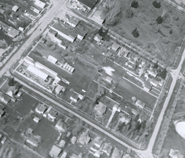 An aerial view of a neighborhood with intersecting streets and rows of small buildings and houses. There is a large open area with pathways and scattered trees, which appears to be a park, occupying part of the scene. The layout shows a mixture of residential and possibly industrial or commercial structures.