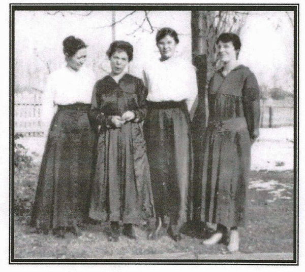 Four women standing in a garden, wearing long skirts and blouses. They are posed side by side with trees and a fence in the background.