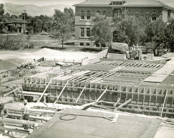 A construction site with wooden framework in the foreground and two workers. A truck and construction equipment are nearby. In the background, there is a large brick building with arched windows, and trees nearby. Houses and mountains can be seen in the distance.