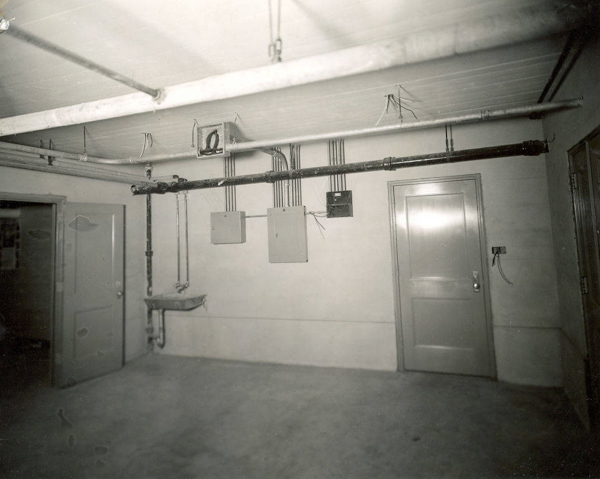 A room with a concrete floor and unfinished walls, featuring two closed wooden doors and one open door. Several pipes and electrical boxes are mounted on the walls. A small sink is attached near the open door, and exposed wiring and pipes run across the ceiling. A light switch with exposed wiring is on the wall near the closed door.