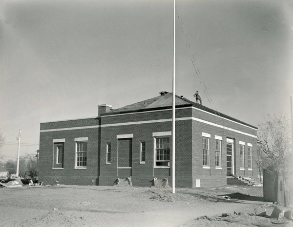 A brick building with large windows and steps leading to an entrance. A person is visible standing on the roof. The surrounding area appears to be a construction site, with dirt and scattered materials. A flagpole is in the foreground.