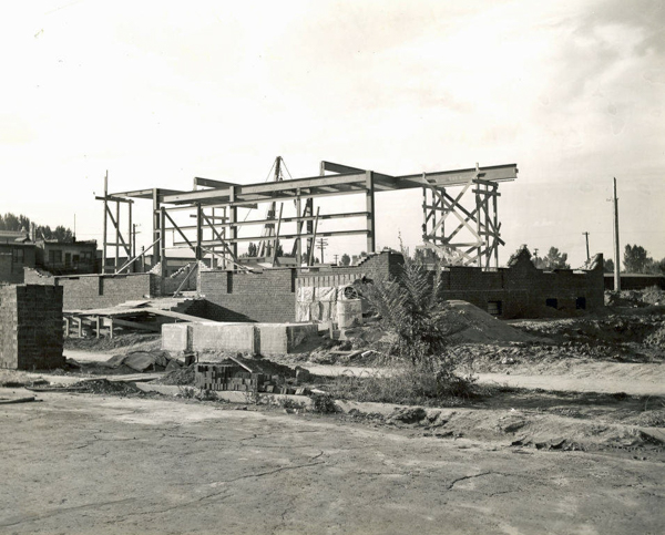 A construction site with a steel framework of a building in progress. Brick walls are partially built, and several stacks of bricks and building materials are scattered around the area. There are utility poles and trees in the background.