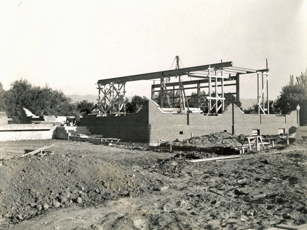 A partially constructed brick building with steel framing and wooden supports. Surrounding the structure are piles of dirt and construction materials. Trees and shrubs are visible in the background.