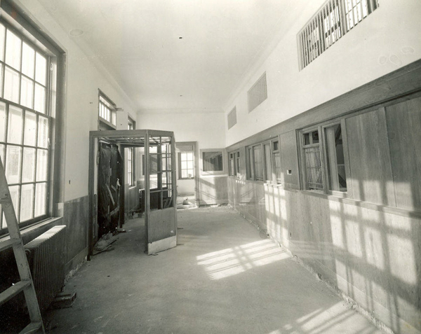 A long hallway with large windows on the left, casting shadows on the floor. There are wooden panels on the right side, interspersed with small barred openings. In the middle of the hallway, there is a partially dismantled phone booth or small enclosure. At the far end, a radiator is visible beneath a window. A ladder leans against the wall on the left.