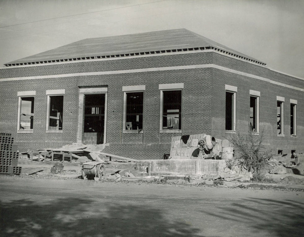 A single-story brick building under construction with several large windows and an open entrance. Construction materials and equipment are scattered around the site, including a cement mixer and stacks of bricks. The ground surrounding the building is unpaved, and there are no trees or landscaping.