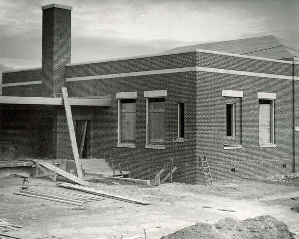 A brick building under construction with several boarded-up windows and a chimney. There is unfinished ground in the foreground with construction materials scattered, including wooden planks and a ladder leaning against the building.