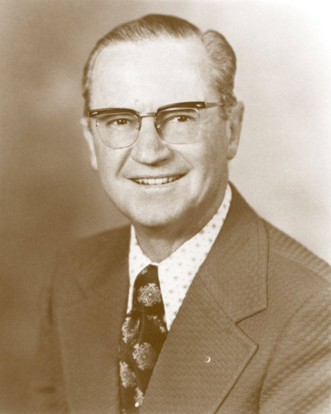 A man with glasses is smiling. He is wearing a patterned suit jacket, a white shirt with small designs, and a tie with floral patterns.