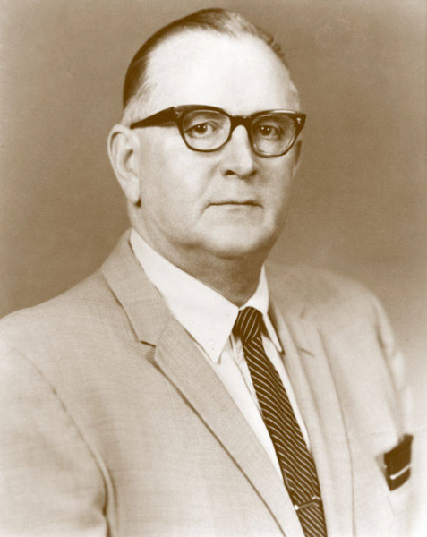 A middle-aged man wearing glasses, a light-colored suit, a white shirt, and a striped tie. His hair is neatly combed back, and he is looking directly at the viewer with a neutral expression.