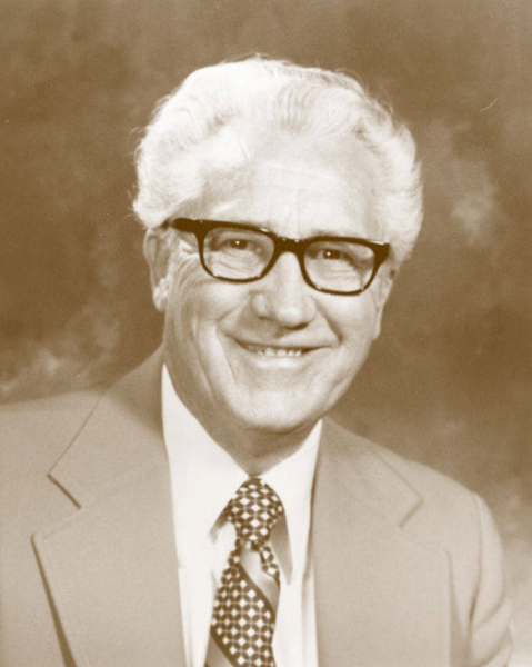 An older man with white hair and glasses is smiling. He is wearing a suit jacket, a collared shirt, and a patterned tie.