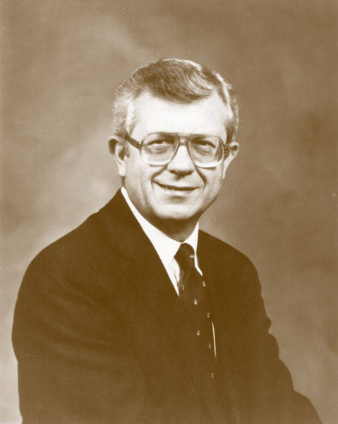 A man wearing glasses and a dark suit with a tie is seated, facing forward with a neutral expression.