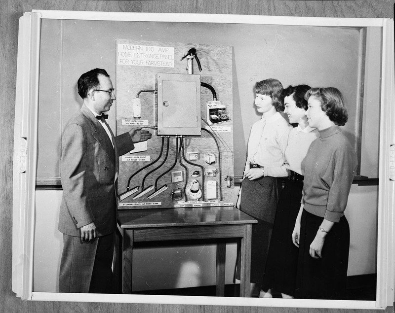 A man showing a labelled electrical panel to three women.
