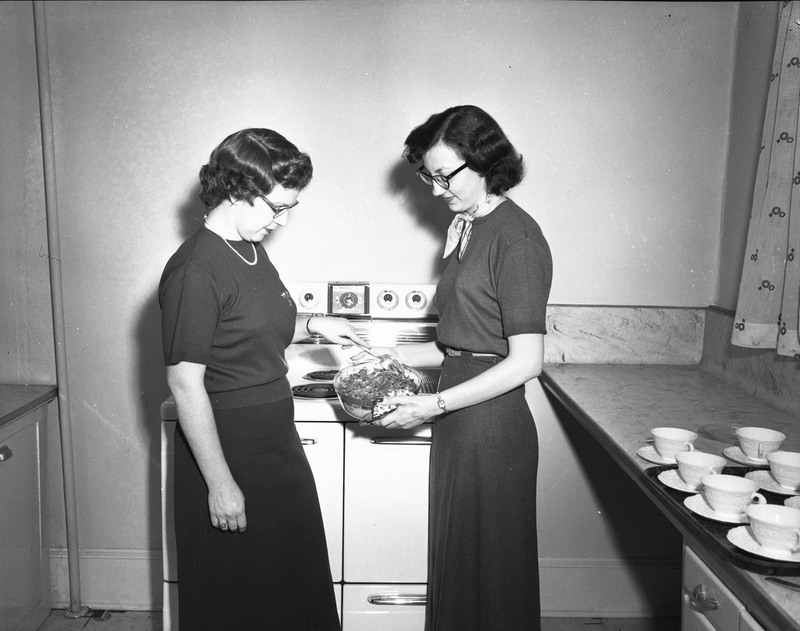Two women preparing food in a kitchen, both looking at a bowl of food.