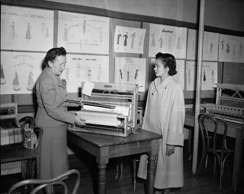 Margret Ritchie (left), Professor of Home Economics at University of Idaho, speaking to foreign student Gertrude Vallejos (right) next to a textile weaving loom. Fashion sewing posters cover the wall behind them.